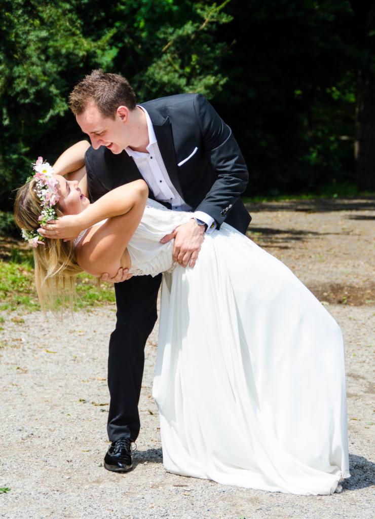 Uśmiechnięta para: mężczyzna w czarnym garniturze podtrzymuje kobietę w białej sukni i wianku z kwiatów, pochyloną do tyłu na żwirowej ścieżce, w tle zielone drzewa.