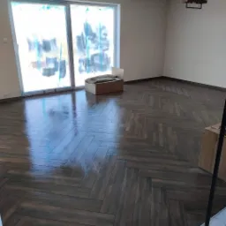 Ułożona w jodełkę, lśniąca, brązowa podłoga z płytek imitujących drewno w pustym pokoju z oknem zabezpieczonym folią malarską.