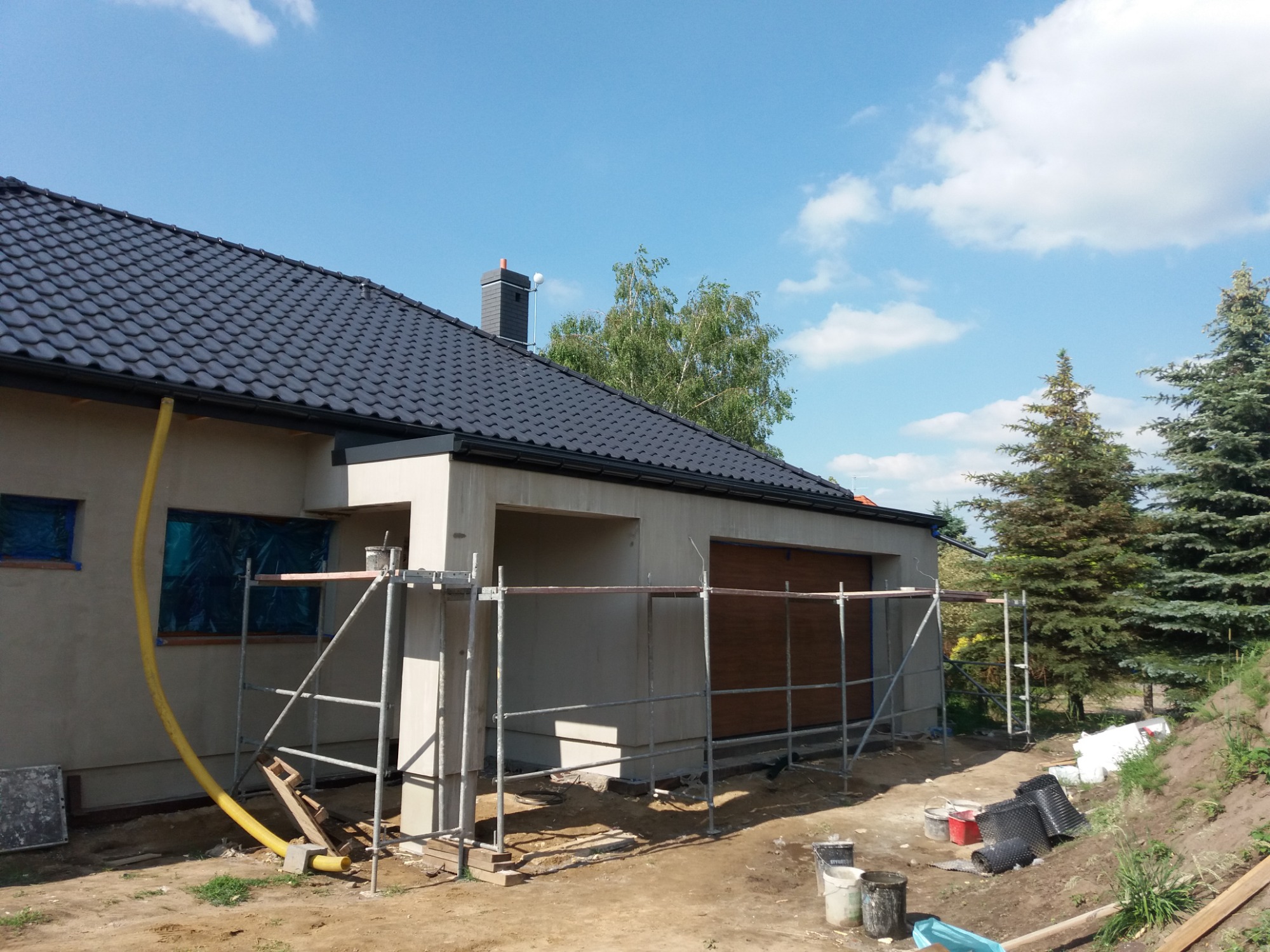 Dom jednorodzinny w trakcie tynkowania elewacji, widoczne rusztowania, zabezpieczone okna folią, czarne dachówki i przygotowane materiały budowlane na ziemi.