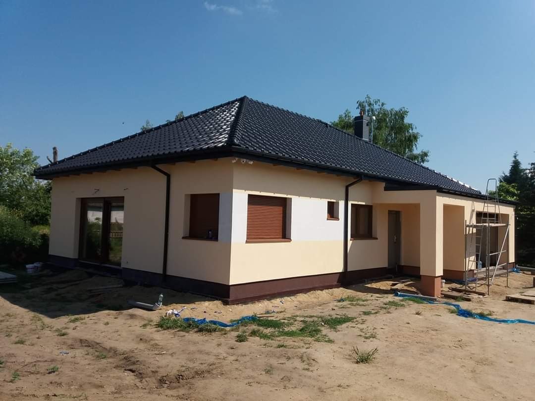 Dom jednorodzinny w Wołczkowie z elewacją w odcieniach beżu i brązu, czarnym dachem i częściowo zasłoniętymi roletami okiennymi; widoczne rusztowanie i teren wokół domu w trakcie prac wykończeniowych.