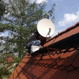 Monter anten satelitarnych na dachu z czerwonej dachówki, zabezpieczony liną, obok białe wiadro, w tle drzewo i błękitne niebo.