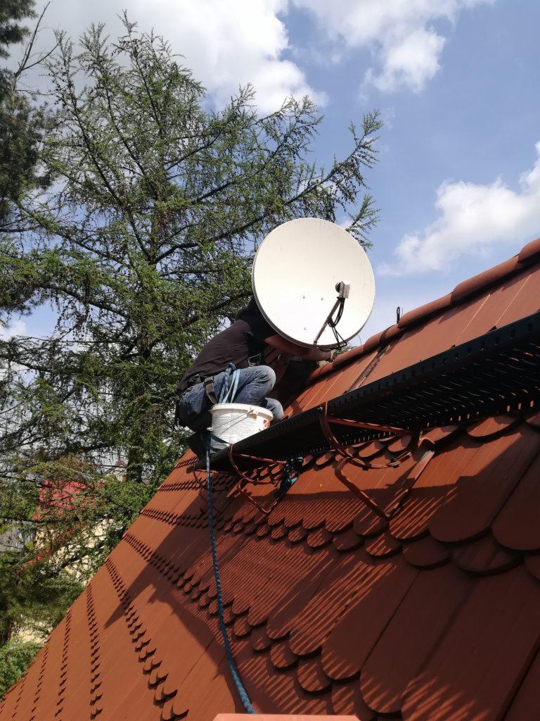 Monter anten satelitarnych na dachu z czerwonej dachówki, zabezpieczony liną, obok białe wiadro, w tle drzewo i błękitne niebo.