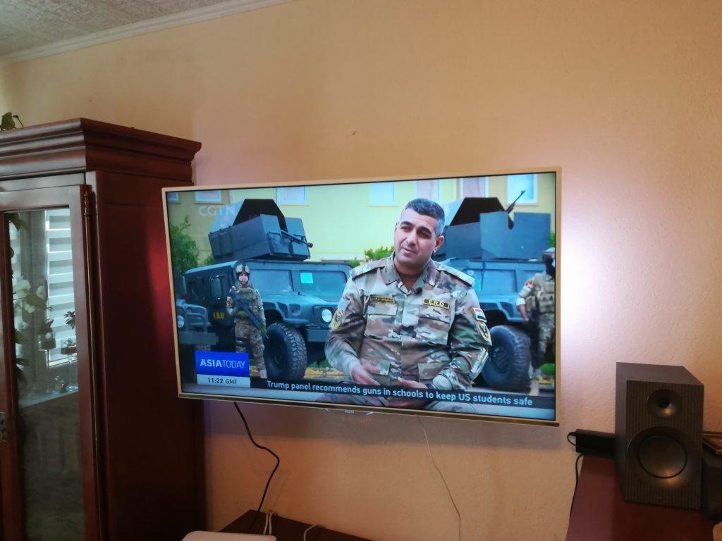 Telewizor LCD zawieszony na ścianie, wyświetlający program informacyjny z żołnierzami i pojazdami wojskowymi w tle, obok głośnik i ciemny drewniany kredens.