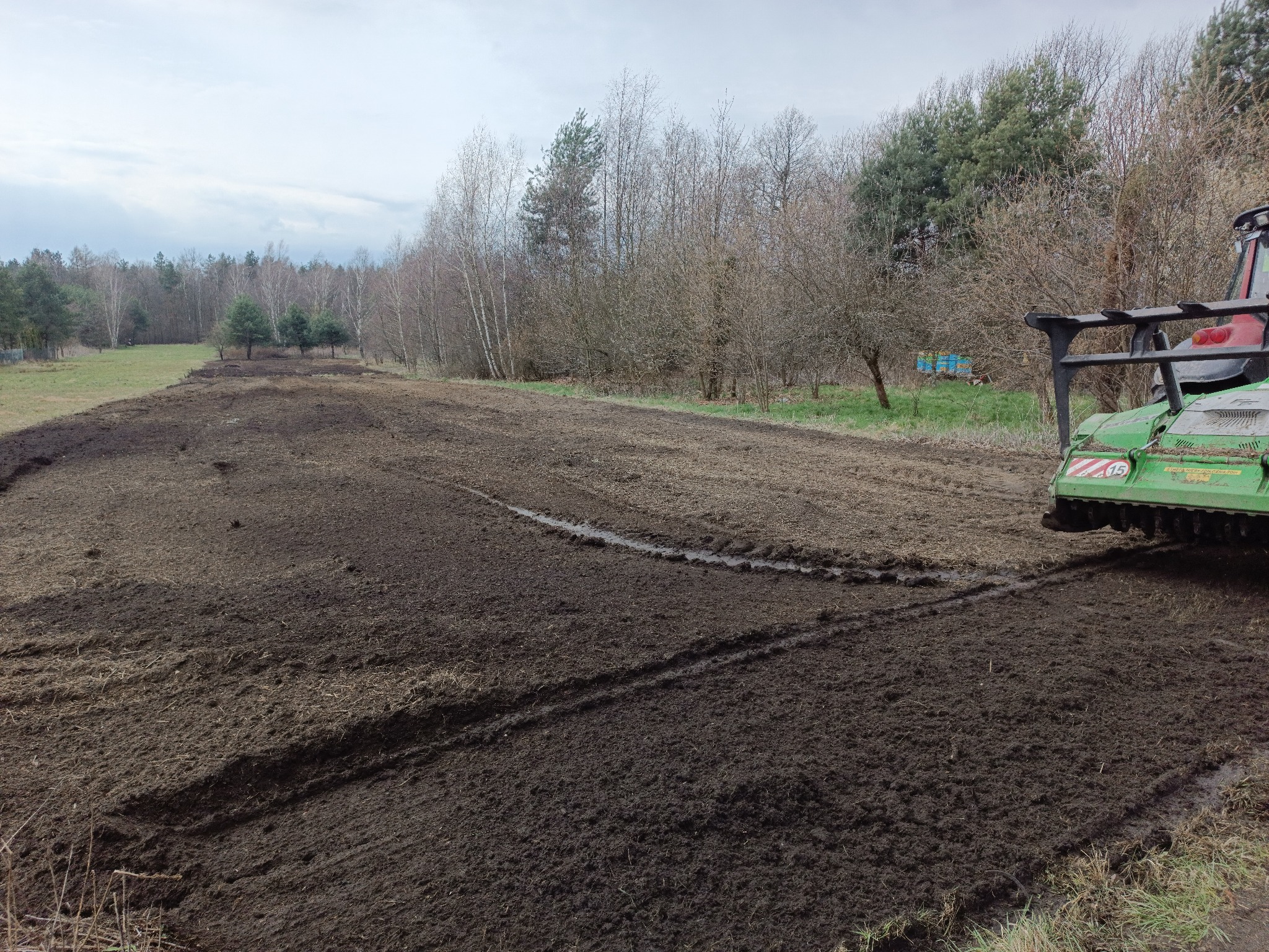 Przygotowane pole pod trawnik z rolki, widoczne ślady po maszynie wyrównującej, w tle las i ul