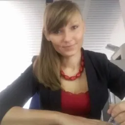 Kobieta w czarnej marynarce i czerwonej bluzce, z czerwonym naszyjnikiem, pisząca długopisem w biurze, z drukarką w tle.