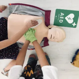 Instruktorzy demonstrują użycie defibrylatora AED na manekinie podczas szkolenia, widoczne zielone rękawice i plansza informacyjna AED.