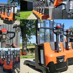 Rząd pomarańczowo-czarnych wózków paletowych elektrycznych Toyota na zewnątrz, z widocznymi panelami sterowania i bateriami, ustawionych na brukowanym placu przed magazynem z panelami słonecznymi...