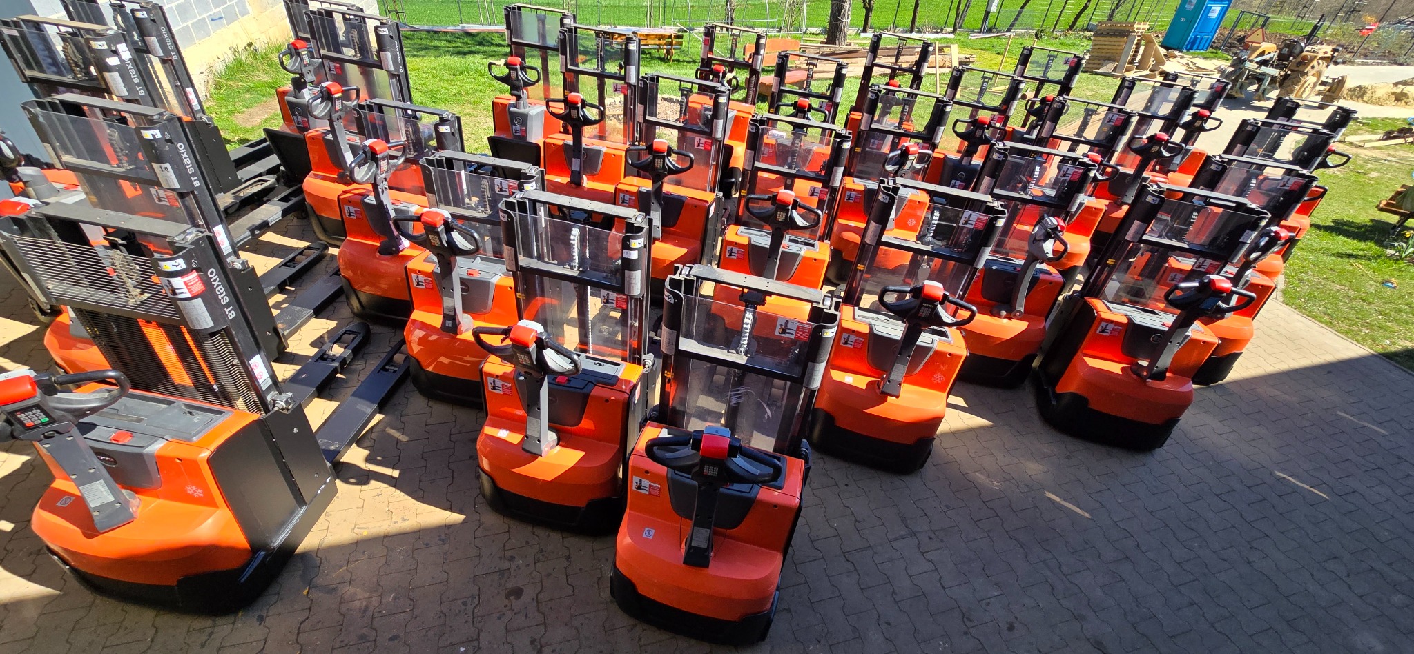 Rząd pomarańczowo-czarnych wózków paletowych elektrycznych ustawionych na brukowanym placu, z widocznym ogrodzeniem i zielenią w tle.