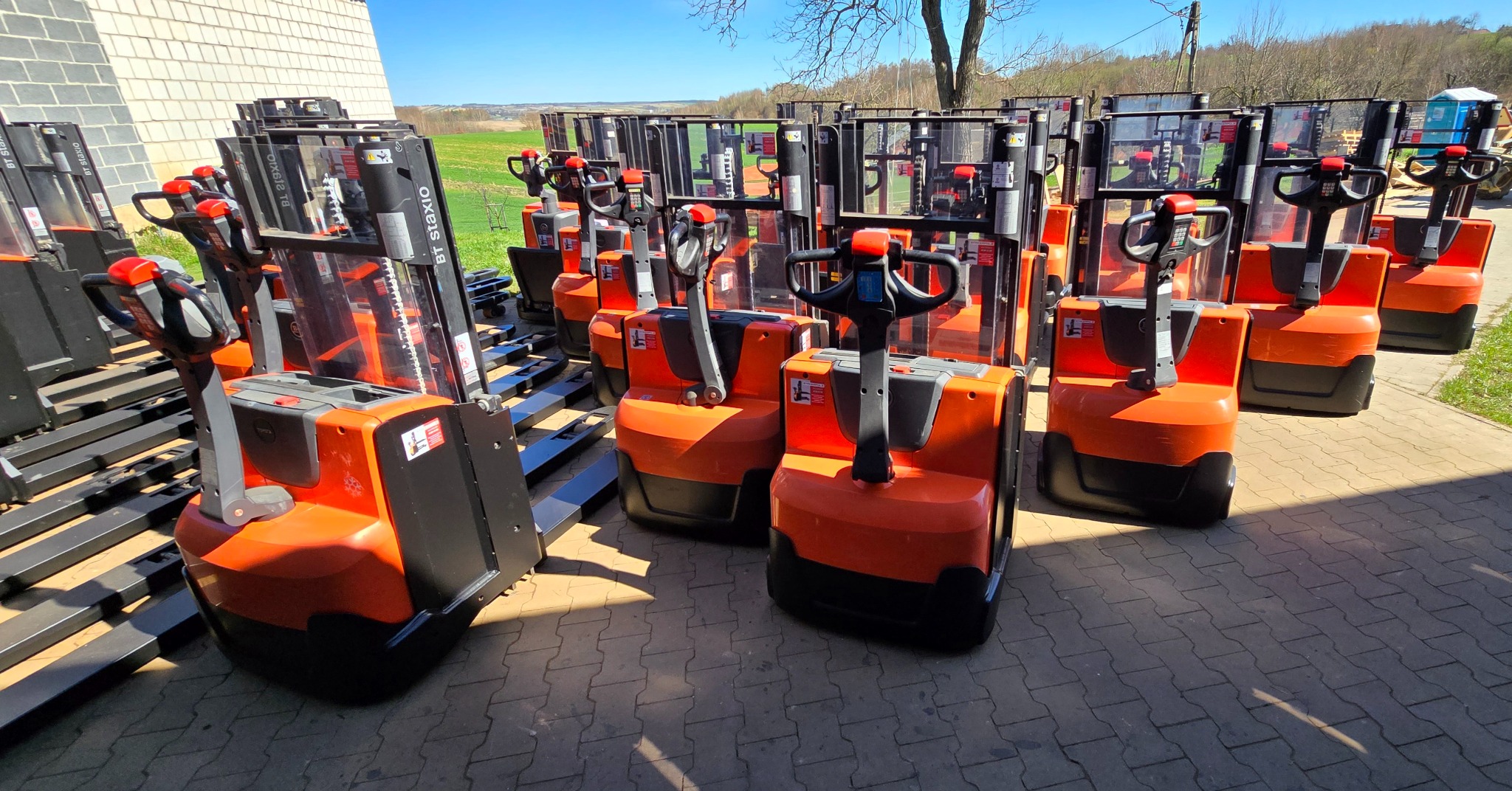 Rząd pomarańczowo-czarnych wózków paletowych z przezroczystymi osłonami, ustawionych na zewnątrz na kostce brukowej, z widokiem na zielone pola i las w tle.