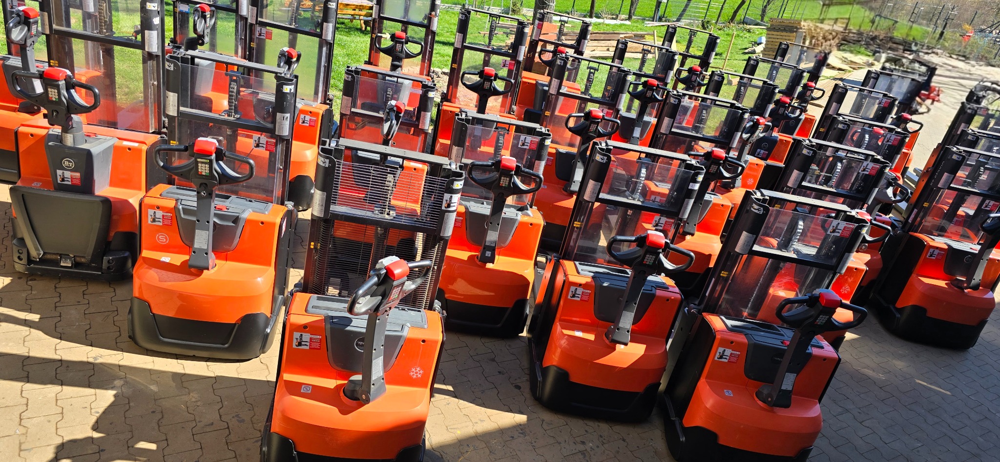 Rząd nowych, pomarańczowo-czarnych wózków paletowych elektrycznych na brukowanym placu, z widoczną trawą i drzewami w tle.