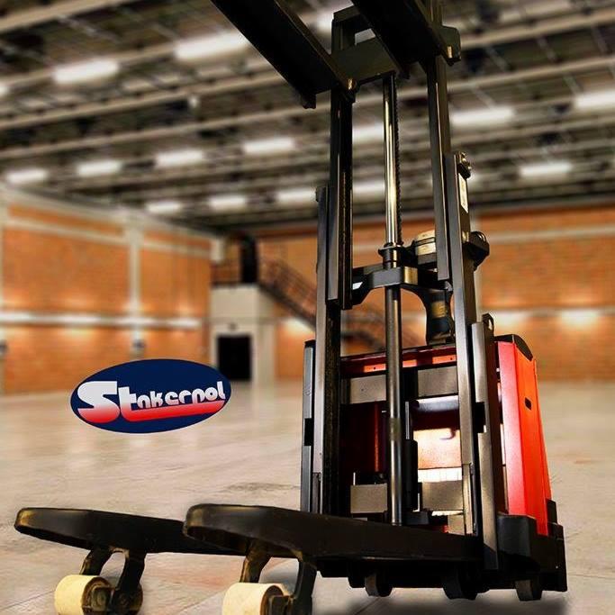 Elektryczny wózek paletowy z logo firmy na tle magazynu z ceglanymi ścianami i schodami.