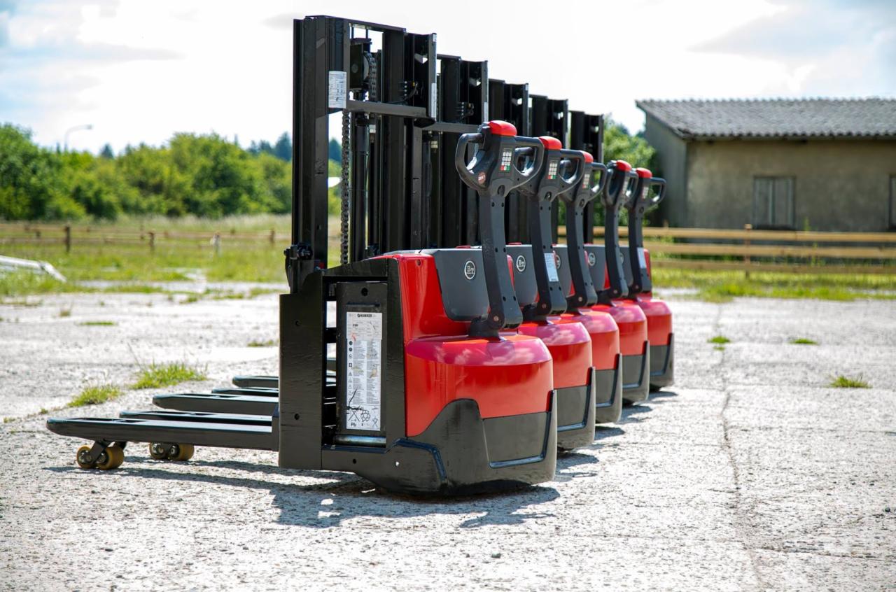 Rząd nowych, czerwono-czarnych wózków paletowych elektrycznych ustawionych na zewnątrz, z widocznymi detalami konstrukcyjnymi i etykietami informacyjnymi.