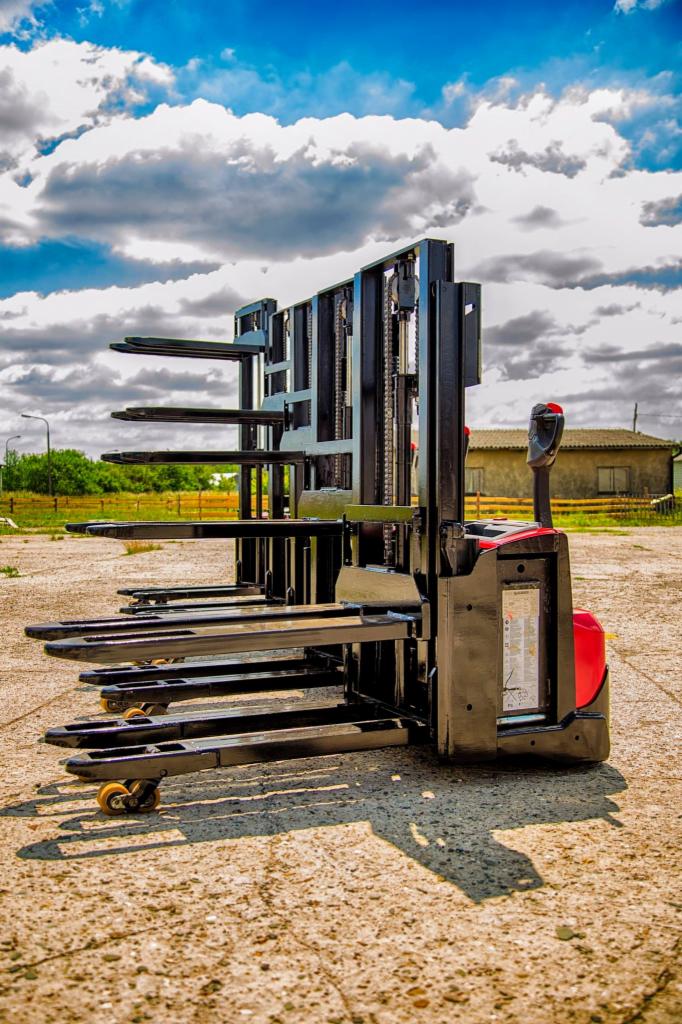 Elektryczny wózek paletowy z wysuniętymi widłami na zewnątrz, sfotografowany na zewnątrz w słoneczny dzień z widocznymi chmurami na niebie.