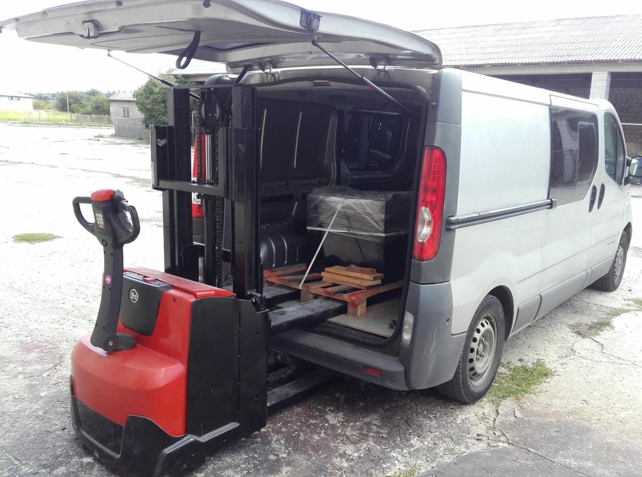 Czerwono-czarny wózek paletowy elektryczny wyładowywany z srebrnego samochodu dostawczego, w tle budynek gospodarczy.