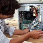 Mężczyzna w szarej koszulce trzyma płytę główną komputera na drewnianym biurku, obok otwarta obudowa komputera stacjonarnego z widocznymi podzespołami.