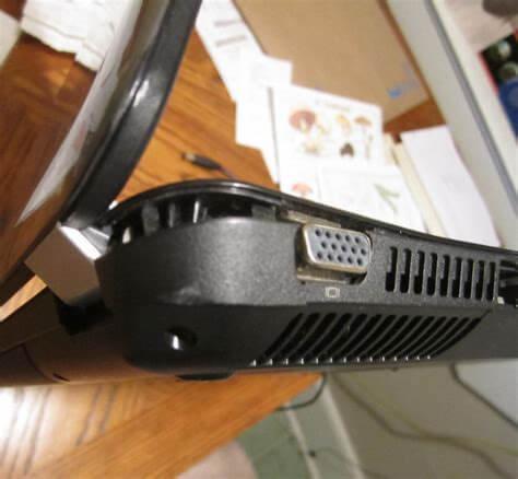 Czarny laptop z widocznym portem VGA i kratką wentylacyjną, leżący na drewnianym stole z rozłożonymi dokumentami w tle.