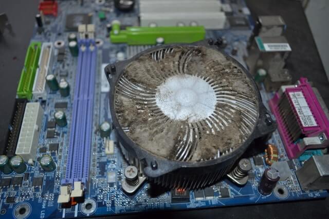 Zabrudzony kurzem wentylator procesora na płycie głównej komputera, widoczne sloty RAM, kondensatory i inne komponenty elektroniczne.