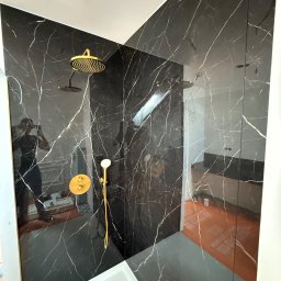 Anton Palianytsia ATOM - BUD - Kabina prysznicowa w łazience wykończonej czarnym marmurem z białymi żyłami. Złota armatura, widoczne odbicie osoby robiącej zdjęcie.