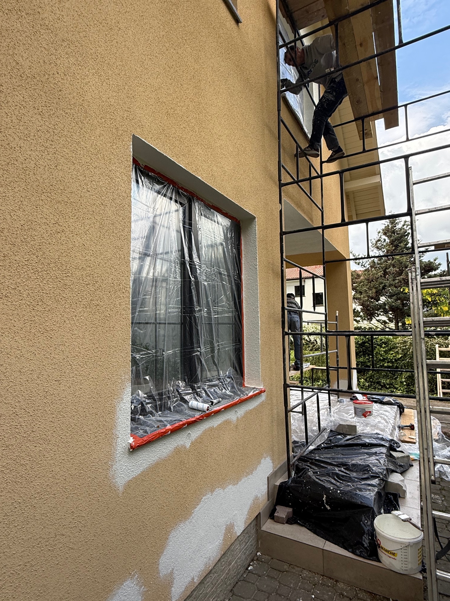 Malowanie elewacji domu z rusztowania. Okno zabezpieczone folią, widoczne ślady farby. Prace w toku, jasna elewacja i drabina.