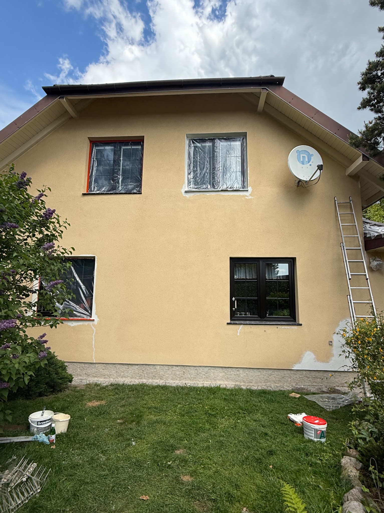 Jasnożółta elewacja domu w trakcie malowania: okna zabezpieczone folią, ślady farby, drabina oparta o ścianę, puszki z farbą na trawie. Słoneczny dzień.