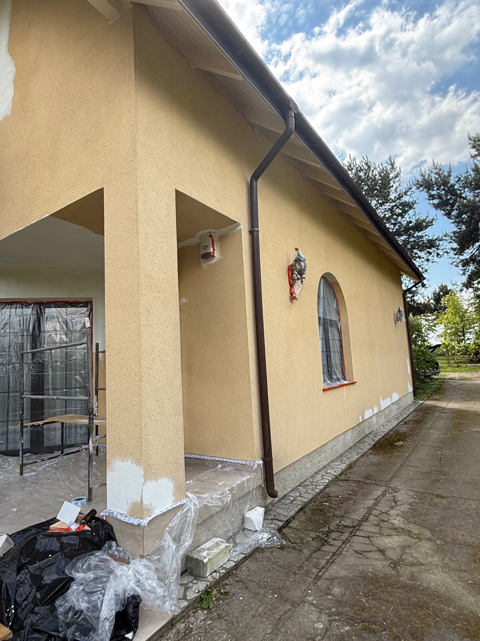 Żółta elewacja domu w trakcie prac wykończeniowych; widoczne zabezpieczone okno, kolumna z fragmentami białej farby, rusztowanie i narzędzia.