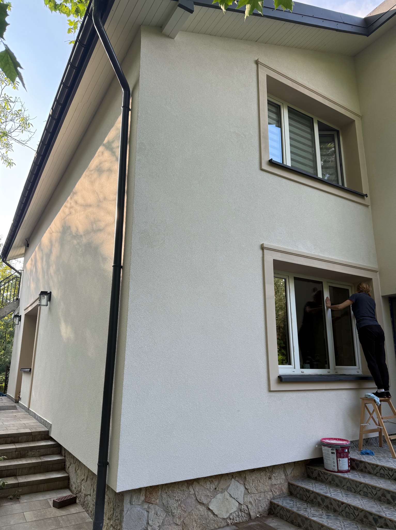 Elewacja domu po renowacji z oknami w beżowych ramach. Kobieta na drabinie czyści okno. Widoczna puszka farby i kamienne wykończenie.