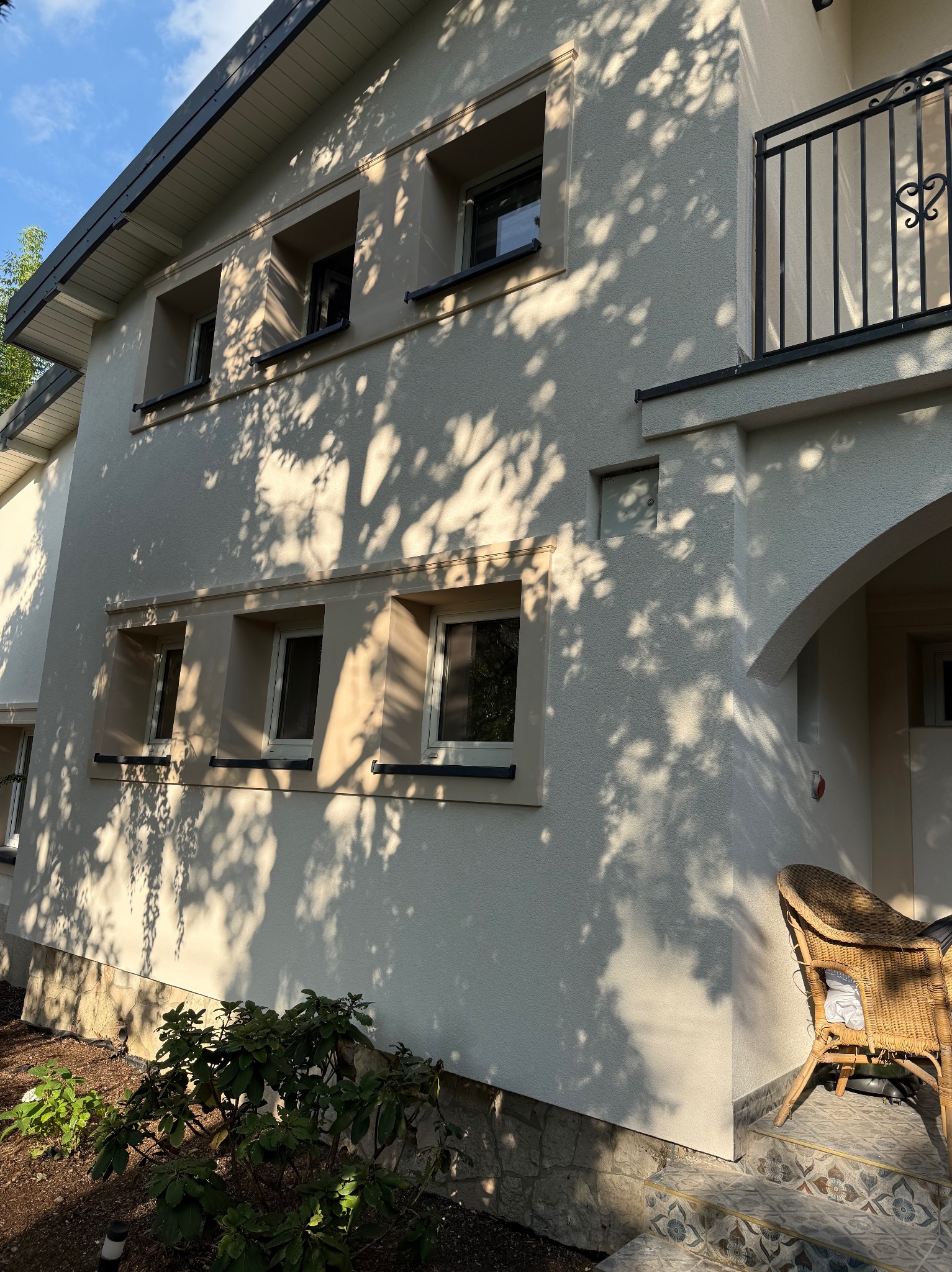 Jasna elewacja domu z balkonem i oknami w ozdobnych ramach, oświetlona przez słońce, z widocznymi cieniami drzew.