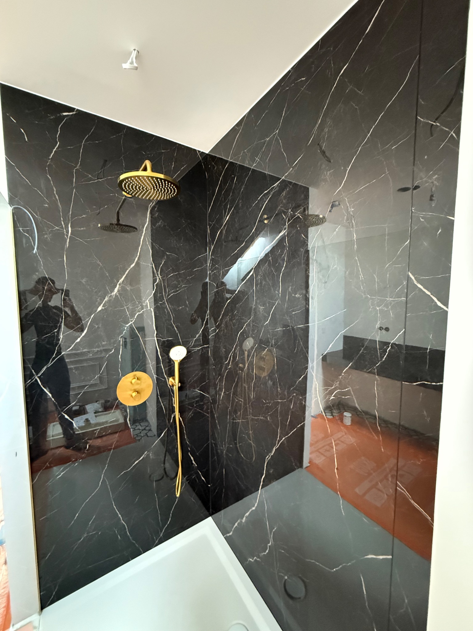 Kabina prysznicowa w łazience wykończonej czarnym marmurem z białymi żyłami. Złota armatura, widoczne odbicie osoby robiącej zdjęcie.