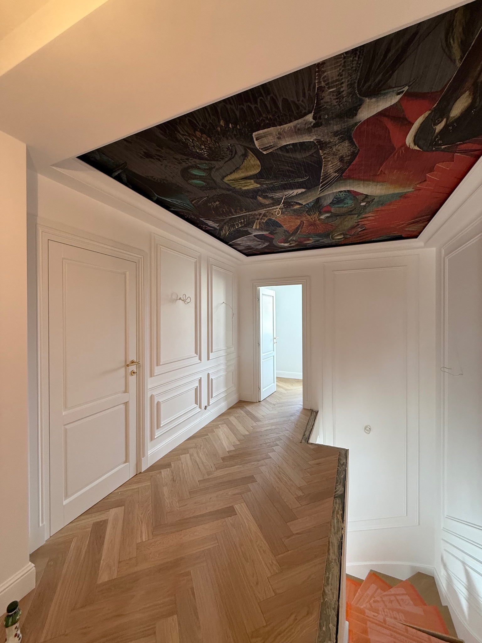 Korytarz po remoncie z parkietem w jodełkę, białą stolarką i ścianami z dekoracyjnymi panelami. Na suficie widoczny mural z motywem ptaków.