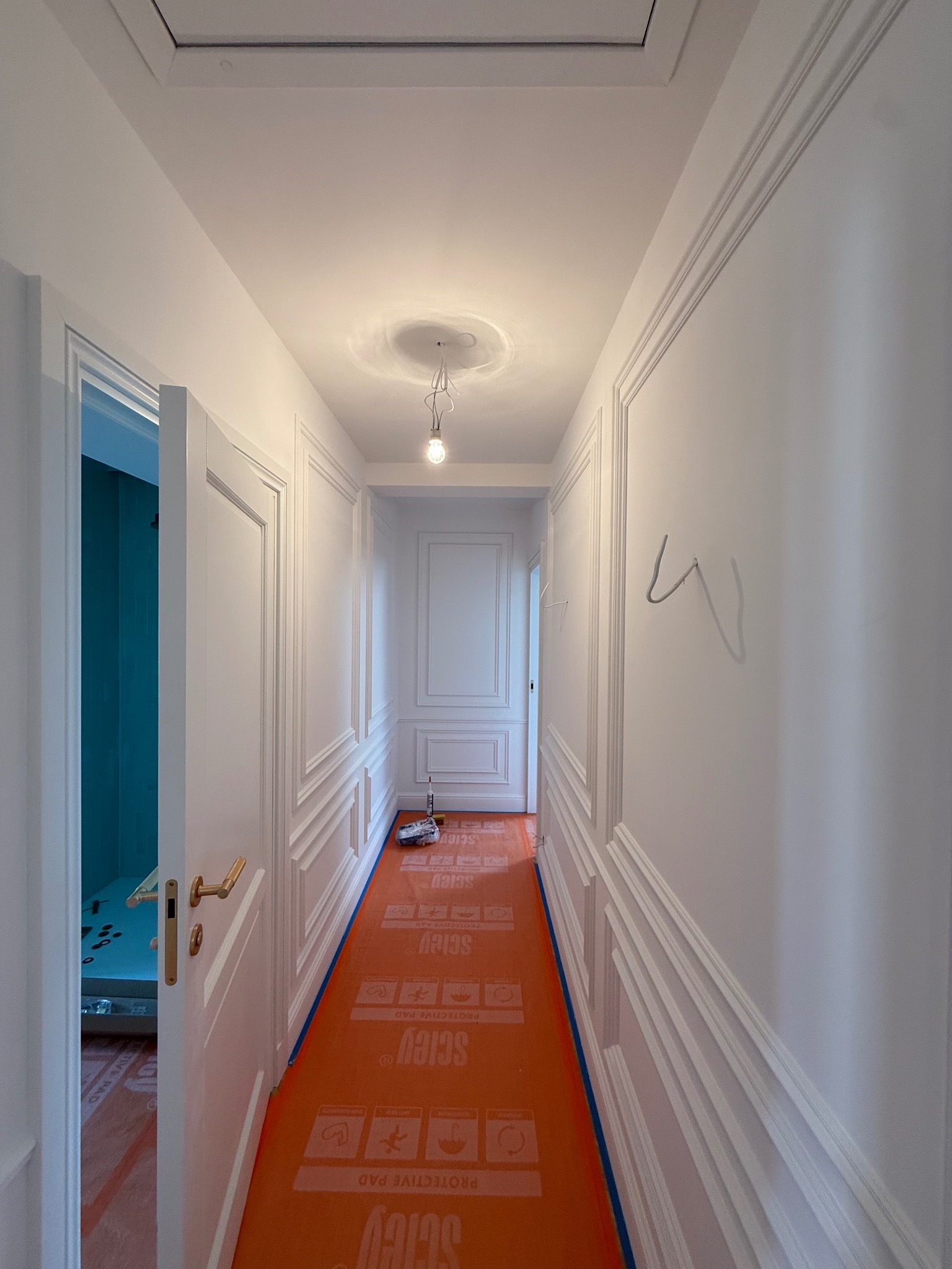 Korytarz po remoncie w Warszawie: białe ściany z dekoracyjnymi listwami, podłoga zabezpieczona pomarańczową folią. Widoczna armatura łazienkowa w tle.