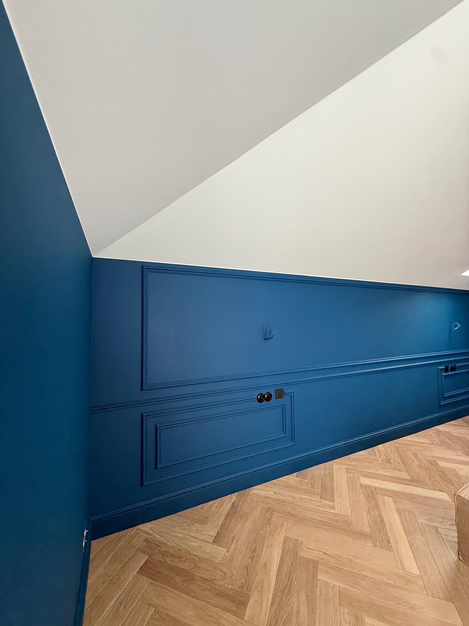 Niebieska ściana z dekoracyjnymi panelami i gniazdkami w wykończonym wnętrzu. Parkiet ułożony w jodełkę. Biały sufit pod skosem.