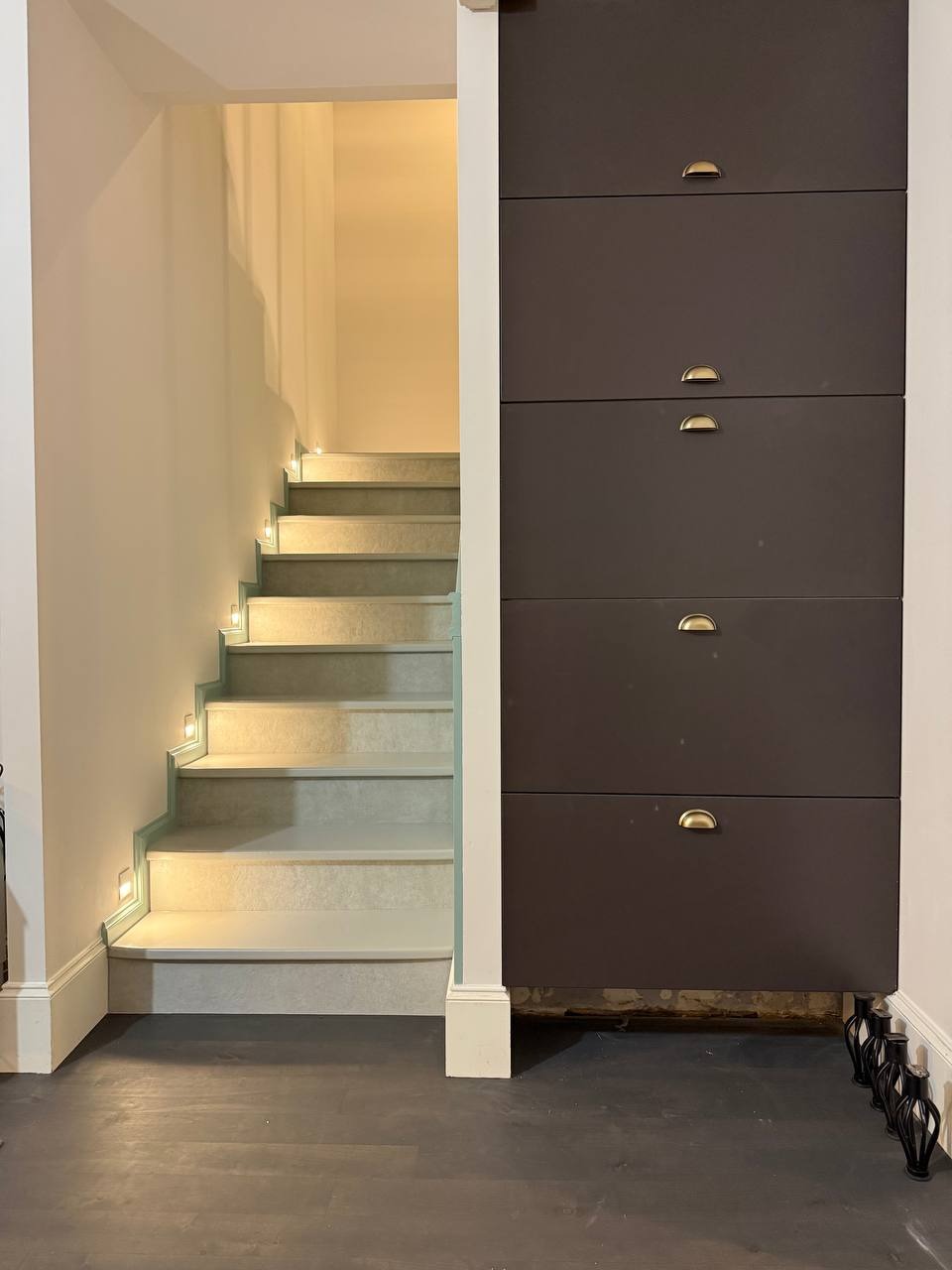 Schody z podświetleniem LED i zabudowa szafkowa w ciemnym kolorze z metalowymi uchwytami, widoczne na tle ciemnej podłogi i jasnych ścian.