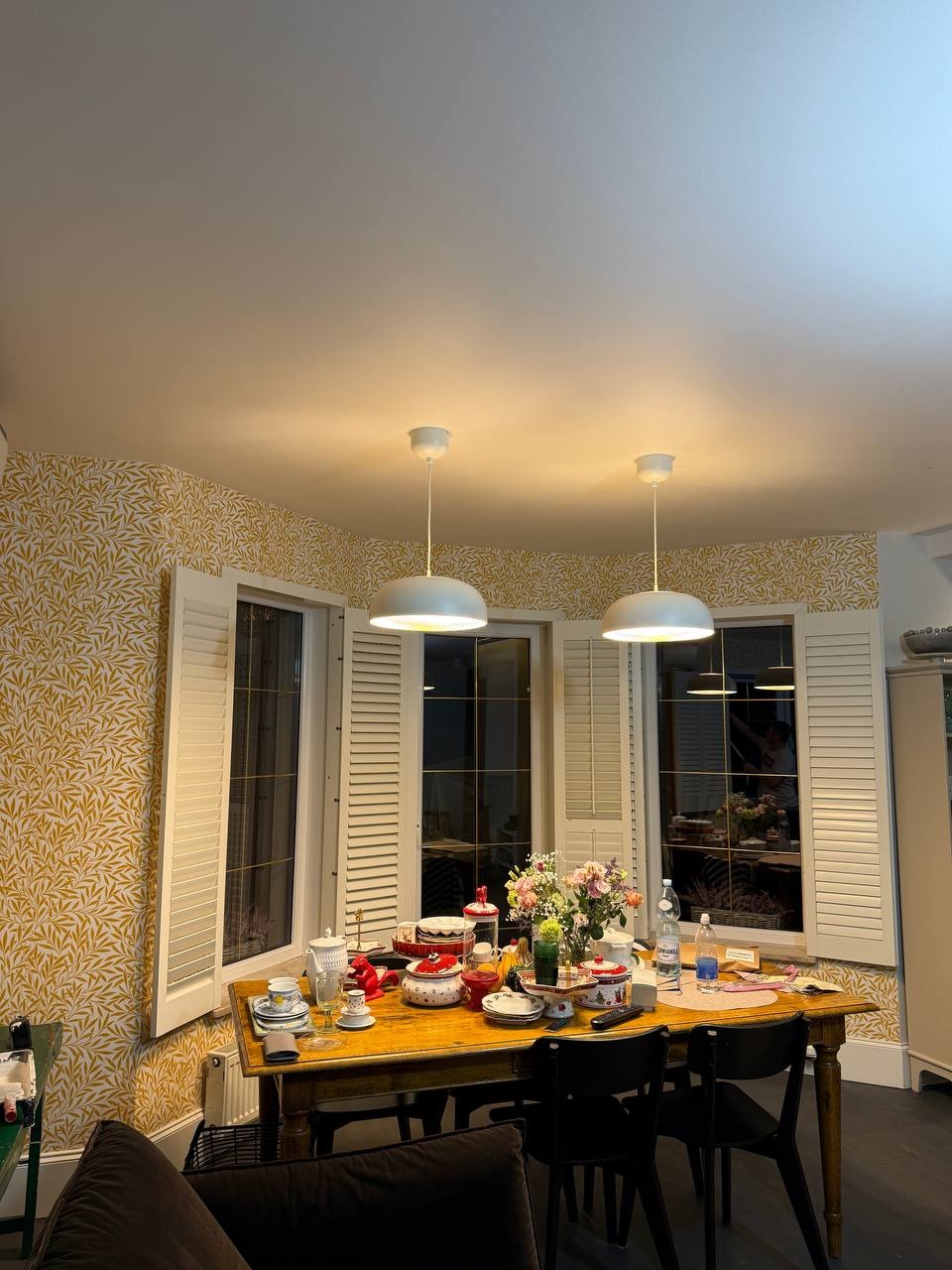 Jadalnia z wzorzystą tapetą w odcieniach złota i bieli, drewnianym stołem zastawionym naczyniami i kwiatami, oświetlona dwoma białymi lampami wiszącymi.