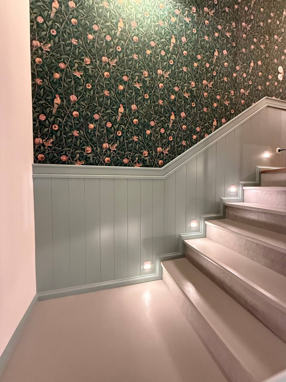 Klatka schodowa po remoncie: wzorzysta tapeta z motywem roślinnym, panele ścienne w odcieniach zieleni i schody z subtelnym oświetleniem punktowym.