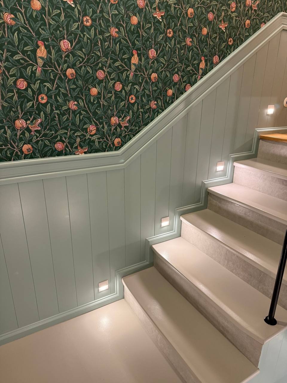 Klatka schodowa po remoncie: pastelowa boazeria z oświetleniem LED, wzorzysta tapeta z motywem roślinnym i odnowione schody w jasnych barwach.