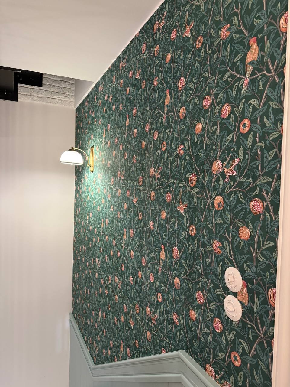 Ściana w zielonej tapecie w stylu vintage z motywem roślinnym i owocowym, oświetlona kinkietem. Widoczne włączniki światła i fragment schodów z jasną balustradą.