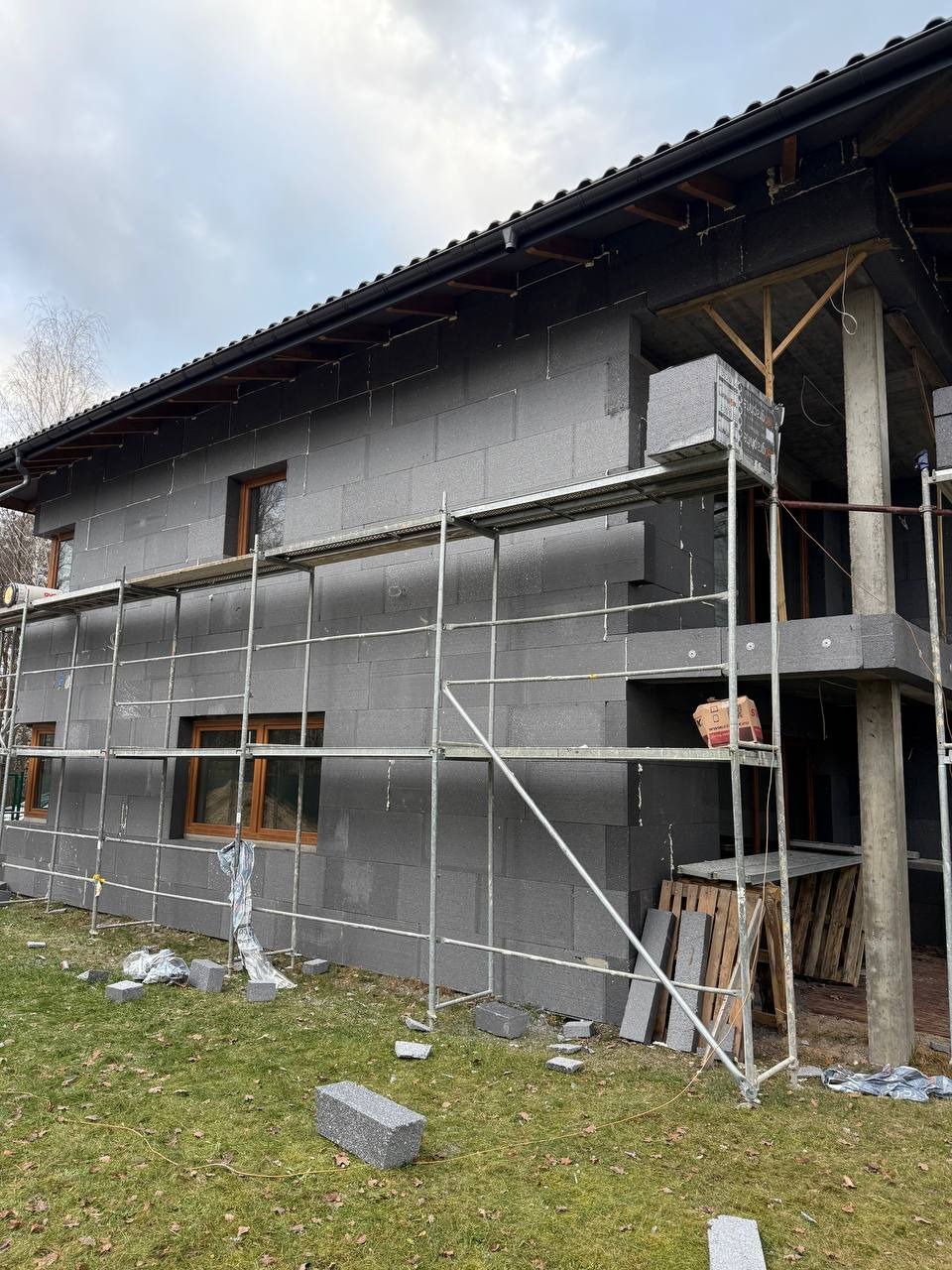 Ocieplanie domu szarym styropianem grafitowym. Widoczne rusztowania, fragmenty styropianu na trawie. Prace w toku, dom w trakcie izolacji termicznej.