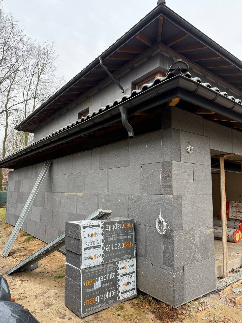 Budynek w trakcie ocieplania szarym styropianem grafitowym. Widoczne pakiety styropianu, rusztowanie i fragment dachu z rynnami. Prace elewacyjne w toku.