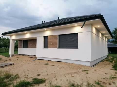 Biały, parterowy dom z czarnym dachem i drewnianymi akcentami okiennymi, oświetlony punktowo, na działce z piaskiem. Widok z boku, pochmurne niebo.