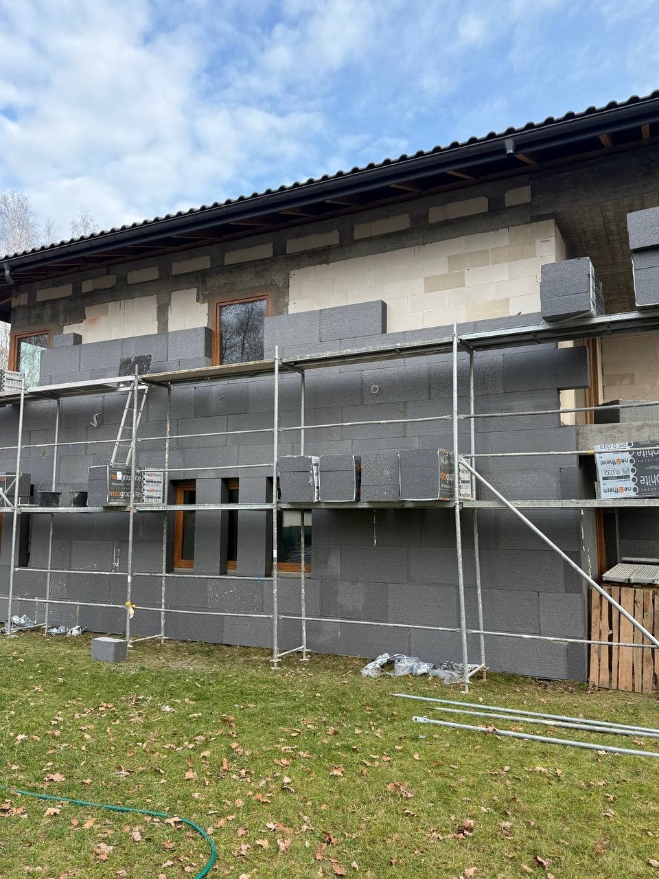 Ocieplanie domu szarym styropianem grafitowym na rusztowaniu. Widoczne okna i fragment dachu. Prace w toku, częściowo ocieplona ściana. Jesienne liście na trawie.