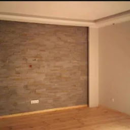 Ściana wewnętrzna wyłożona dekoracyjną okładziną z kamienia w odcieniach beżu i szarości, z widocznymi gniazdkami elektrycznymi i puszką instalacyjną, w nowym pomieszczeniu z drewnianą podłogą...