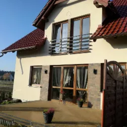 Jasna elewacja domu jednorodzinnego z brązową dachówką i drewnianymi oknami, widok z ogrodu na taras z dekoracyjną drewnianą bramką.