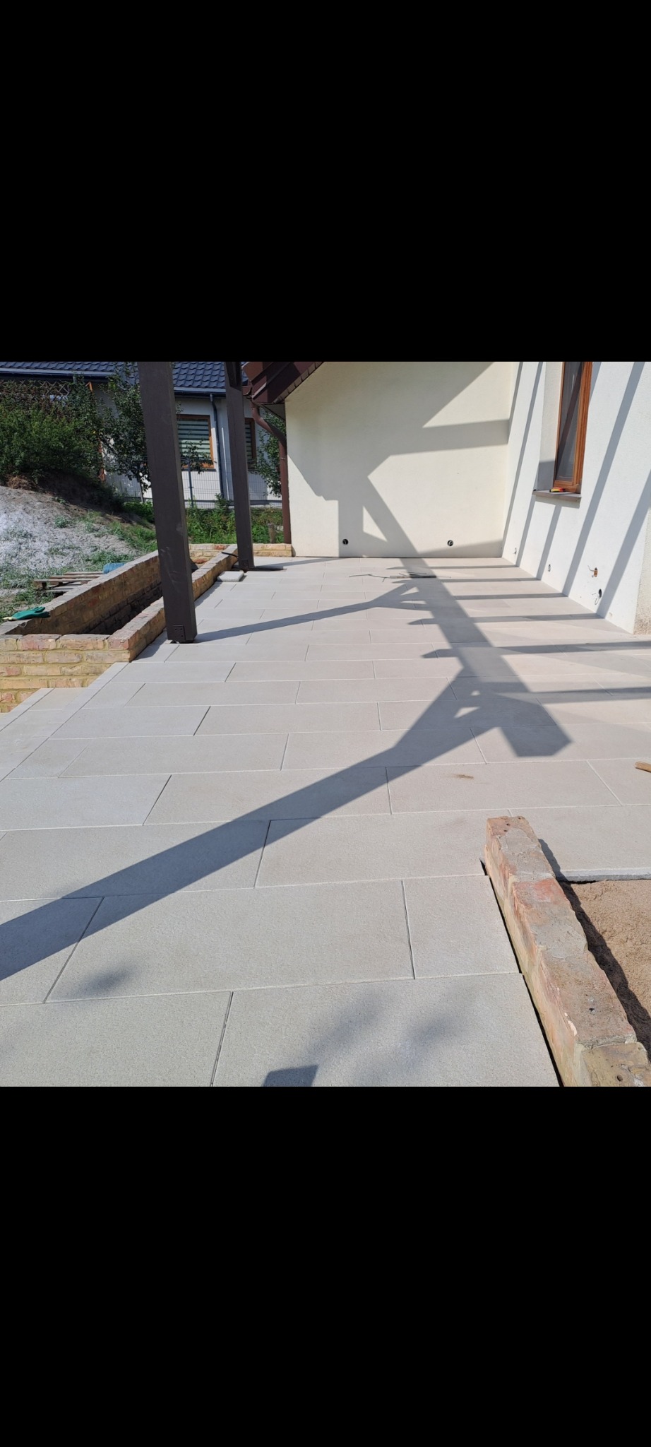 Jasnoszara kamienna posadzka tarasu w trakcie budowy, z widocznymi cieniami pergoli, fragmentem ceglanego obrzeża i przygotowanym podłożem.