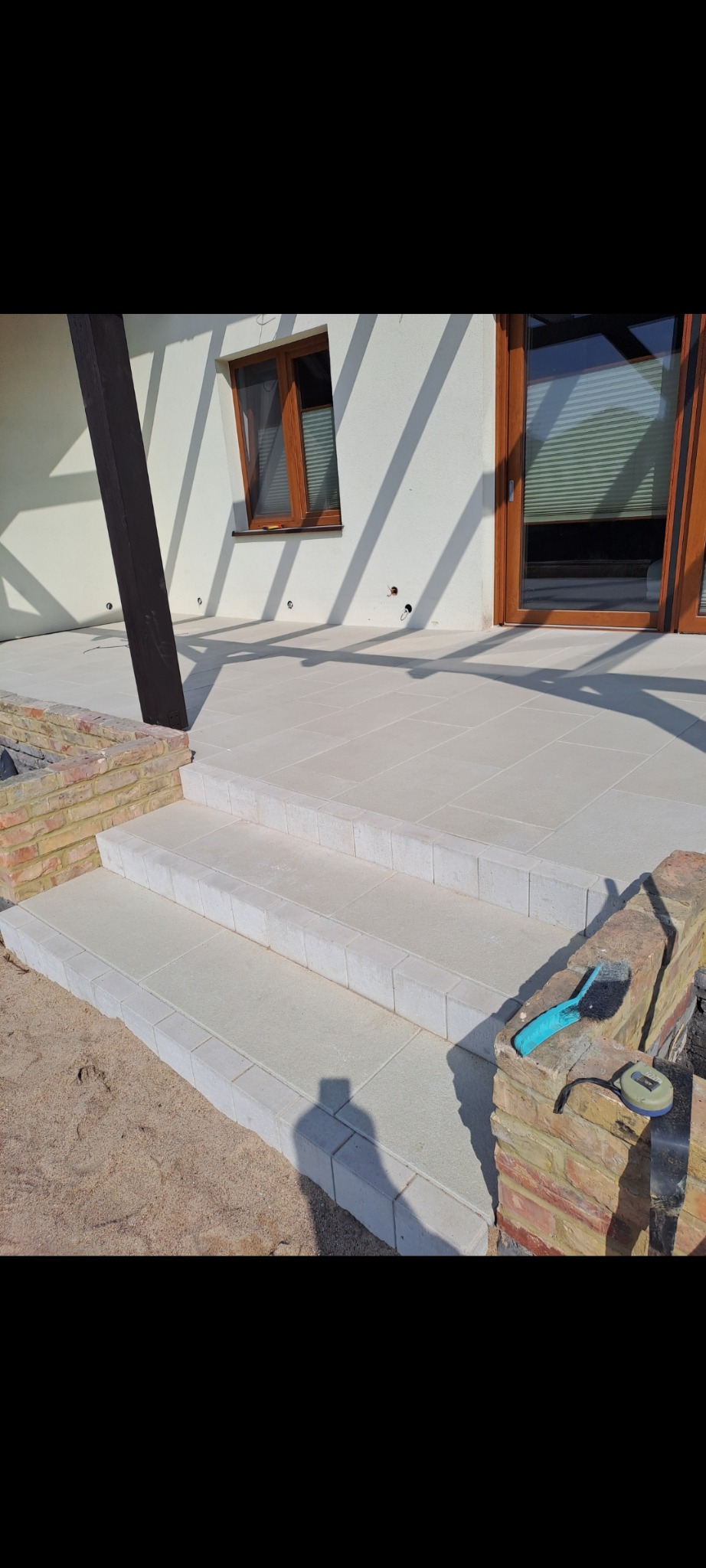 Jasne, kamienne schody prowadzące na taras, z widocznymi narzędziami budowlanymi i cieniem osoby fotografującej.