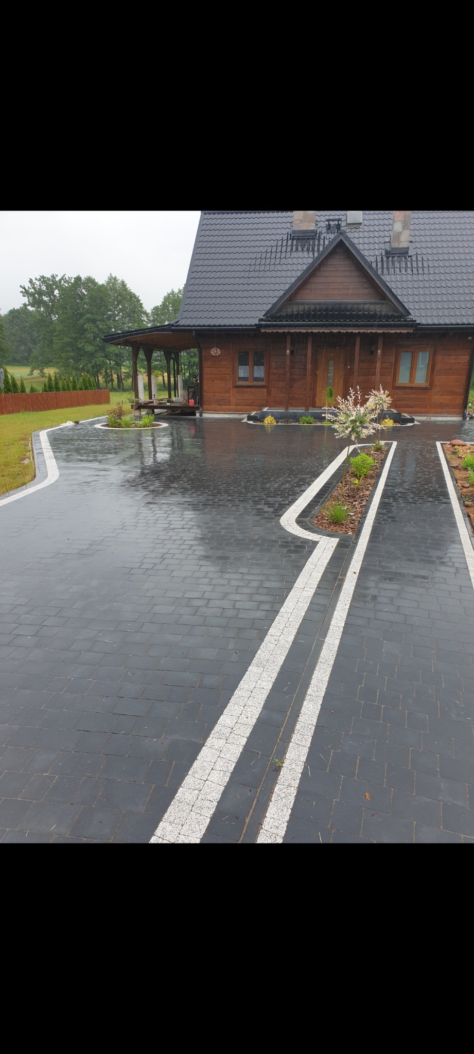 Ułożona ciemnoszara kostka brukowa z białymi akcentami na posesji z drewnianym domem w tle, widoczne rabaty z roślinami i drzewkami, mokra nawierzchnia po deszczu.