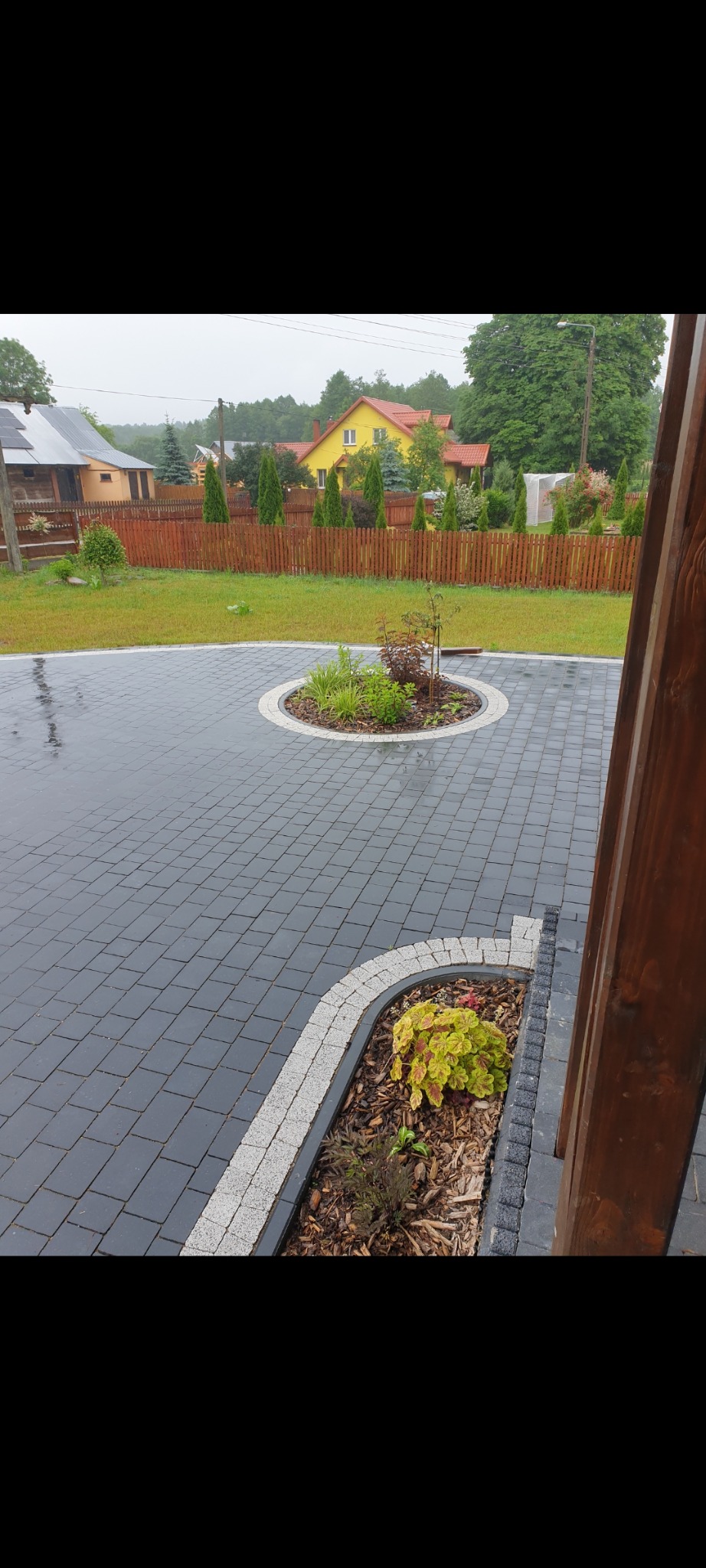 Ułożona szara kostka brukowa z okrągłą rabatą pośrodku i małą rabatą przy drewnianym słupku, widoczne budynki w tle.