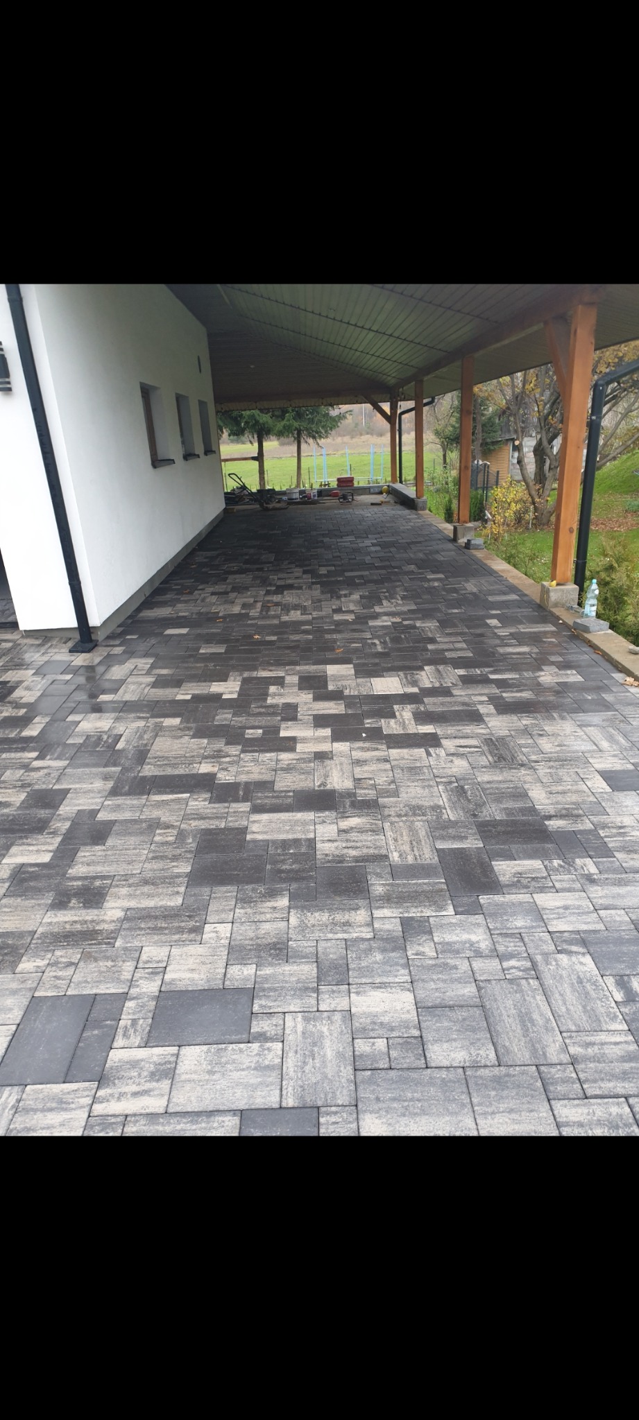 Ułożona, szaro-biała kostka brukowa na podjeździe z zadaszeniem, widok z góry, na obrzeżach widoczna butelka wody i narzędzia ogrodnicze, w tle trawnik i drzewa.