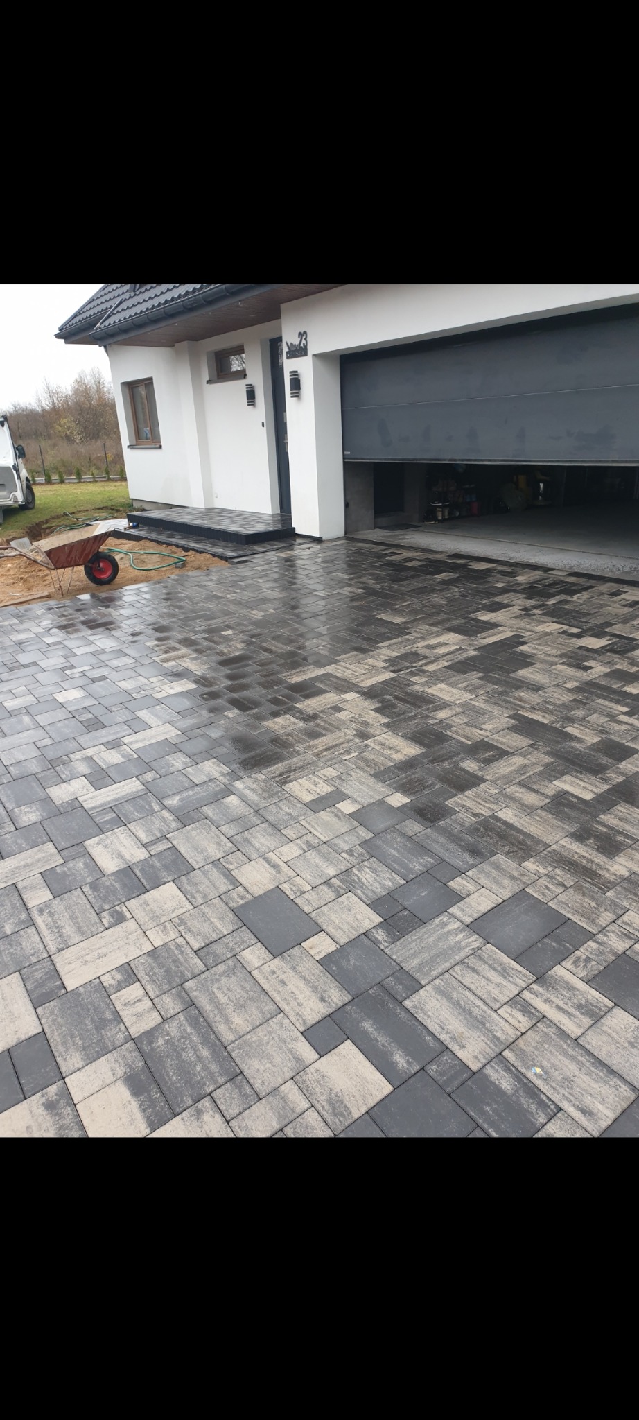 Ułożona kostka brukowa w odcieniach szarości prowadząca do garażu i wejścia do domu z numerem 23, widoczny wózek budowlany z czerwoną felgą.