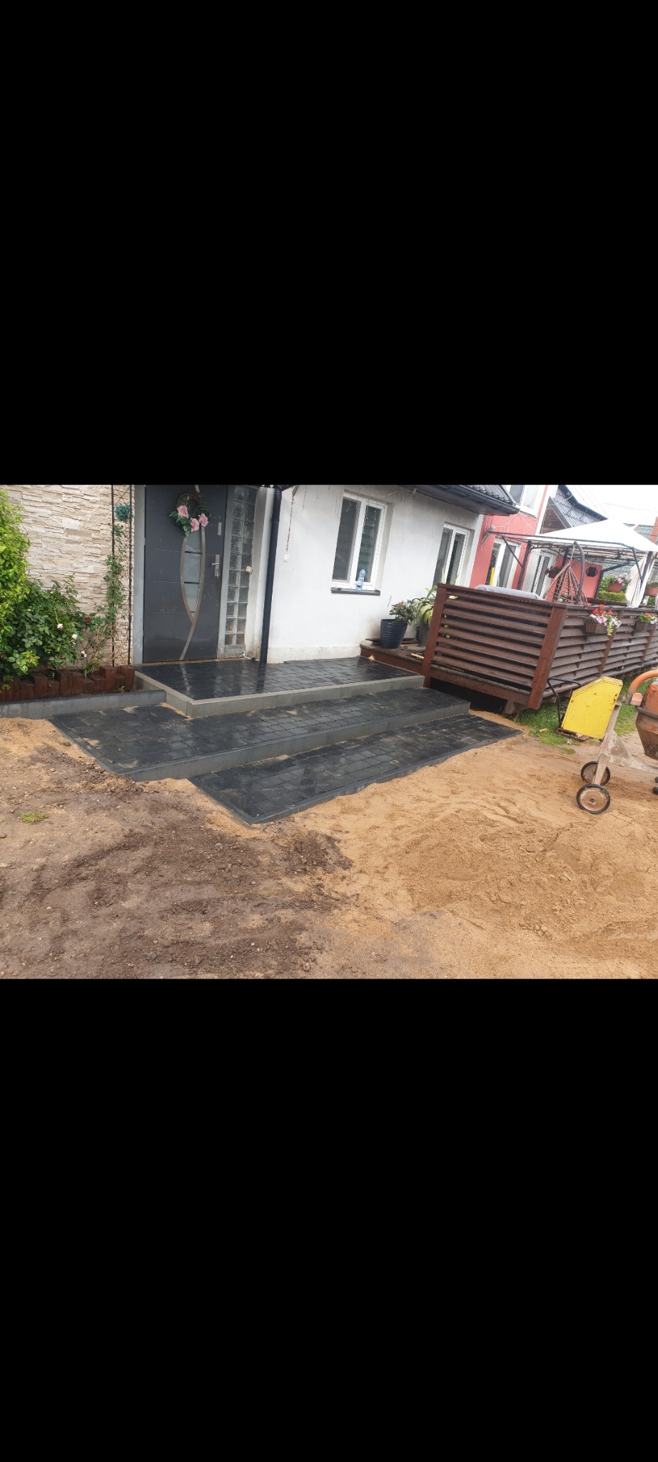 Ułożona ciemna kostka brukowa prowadząca do wejścia domu z drewnianymi schodami i dekoracjami kwiatowymi, widoczny piasek i betoniarka.