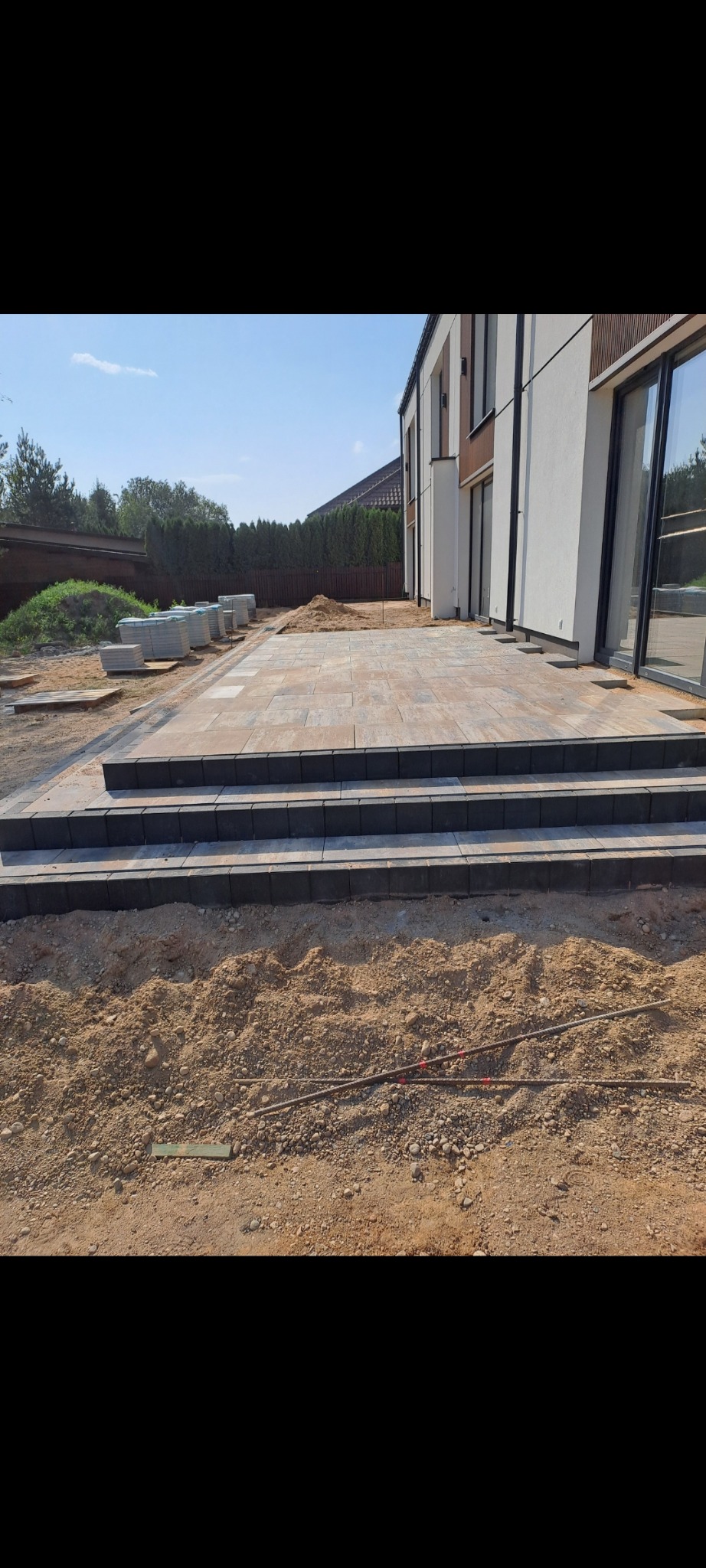 Nowoczesny taras z kamienną posadzką w odcieniach beżu i brązu, z trzema stopniami wykończonymi ciemnoszarą kostką, w tle biała elewacja budynku z dużymi oknami, widoczne przygotowane kostki...